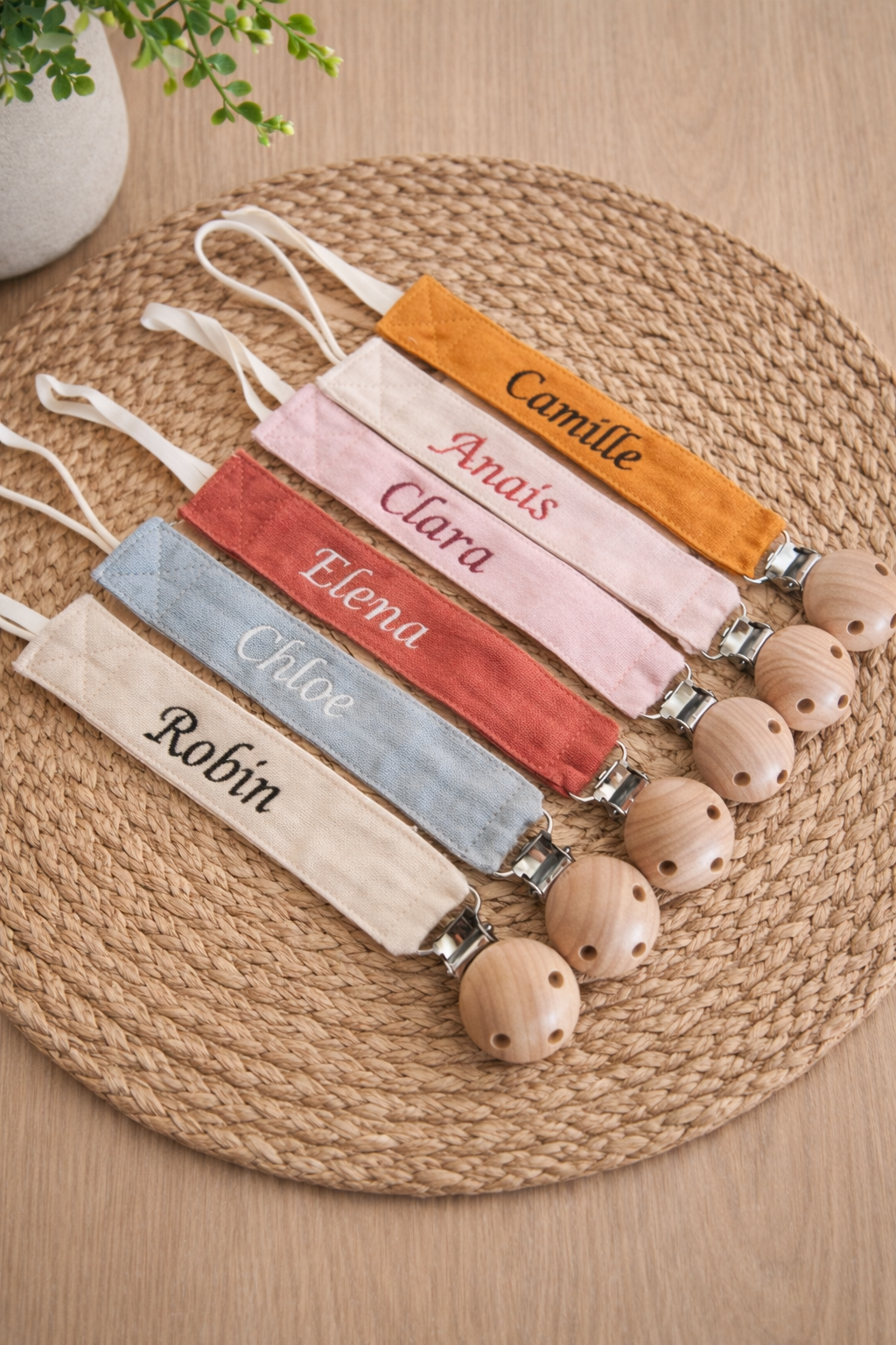 Lot d’attaches-sucettes en tissu de différentes couleurs (beige, bleu, rouge, rose et moutarde) avec prénoms brodés, posées sur un set de table en fibres naturelles, avec clips en bois.