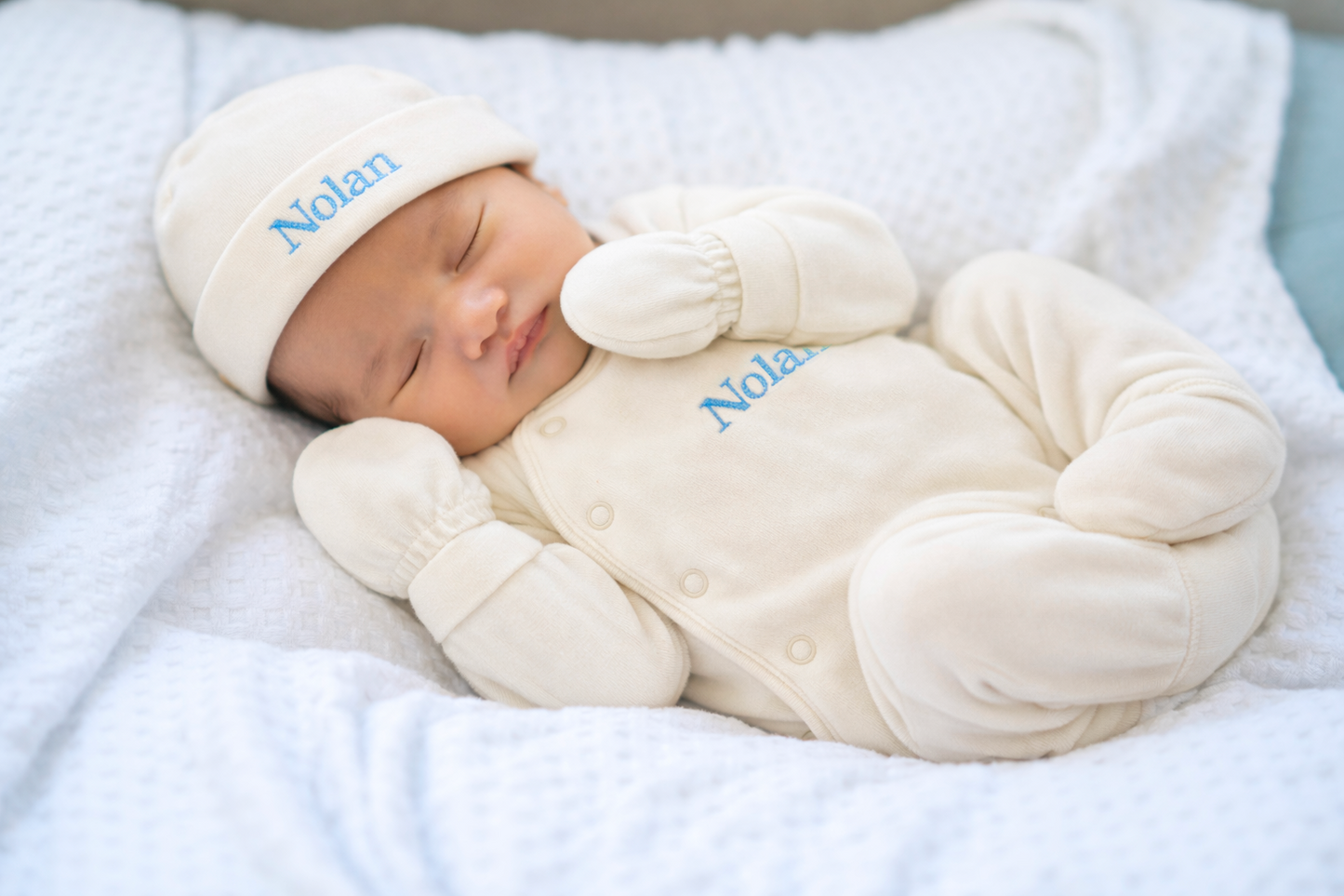 Nouveau-né endormi sur une couverture douce, habillé d’un pyjama beige avec le prénom “Nolan” brodé en bleu, portant également un bonnet et des moufles assortis.