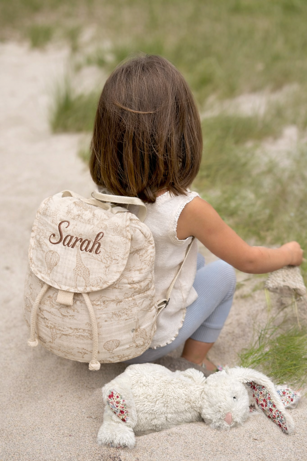 Enfant portant un sac à dos bébé personnalisé avec prénom brodé, motif savane beige, assise en extérieur avec doudou