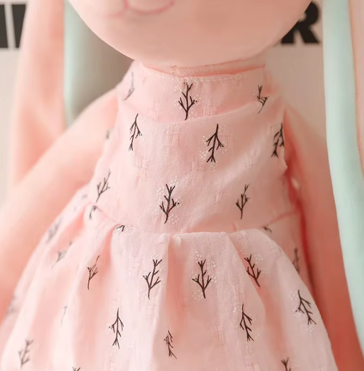 Peluche en forme de lapin rose portant une robe, avec de longues oreilles et un nœud sur la tête, assise sur fond blanc.