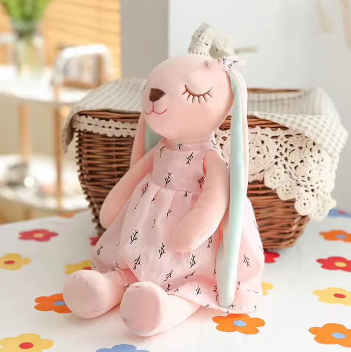 Peluche en forme de lapin rose portant une robe, avec de longues oreilles et un nœud sur la tête, assise sur fond blanc. panier 