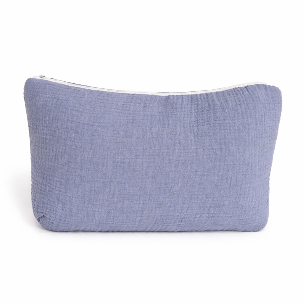 Trousse de toilette rectangulaire en gaze de coton bleu avec le prénom Gabriel brodé en bleu foncé, posée devant des trousses rose et beige avec décor en rotin en arrière-plan.