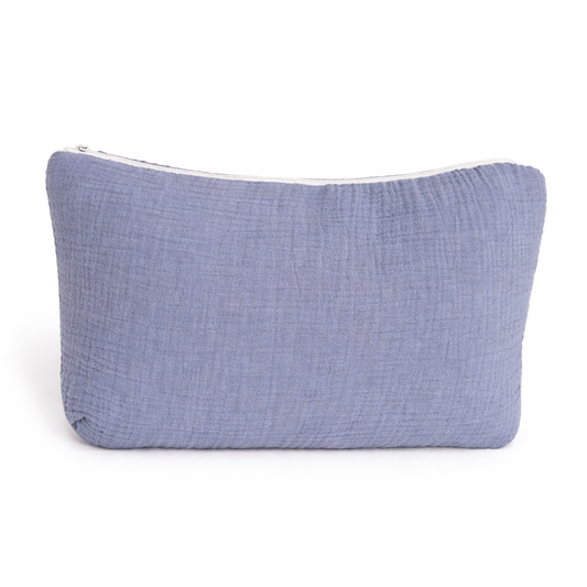 Trousse de toilette rectangulaire en gaze de coton bleu avec le prénom Gabriel brodé en bleu foncé, posée devant des trousses rose et beige avec décor en rotin en arrière-plan.