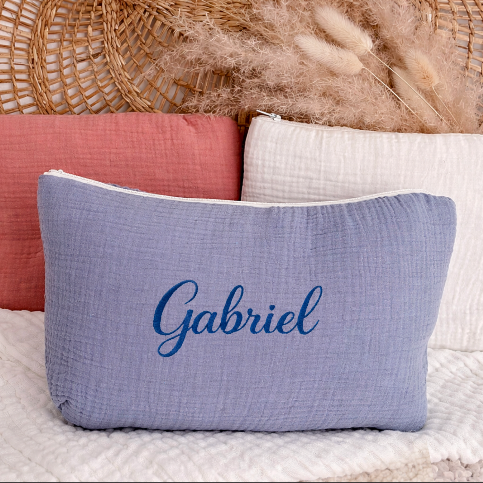Trousse rectangulaire en gaze de coton bleu avec le prénom Gabriel brodé en bleu foncé, posée devant des trousses rose et beige avec décor en rotin en arrière-plan.