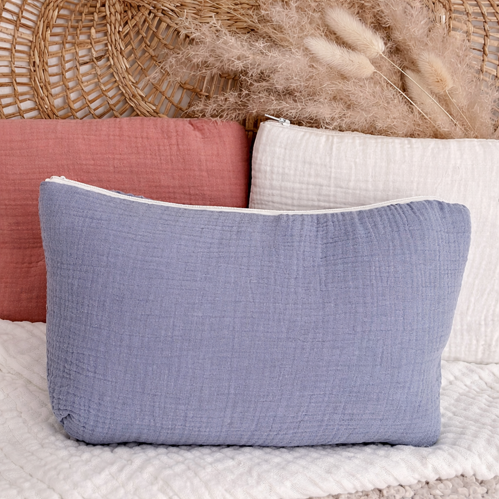 Trousse rectangulaire en gaze de coton bleu avec le prénom Gabriel brodé en bleu foncé, posée devant des trousses rose et beige avec décor en rotin en arrière-plan.