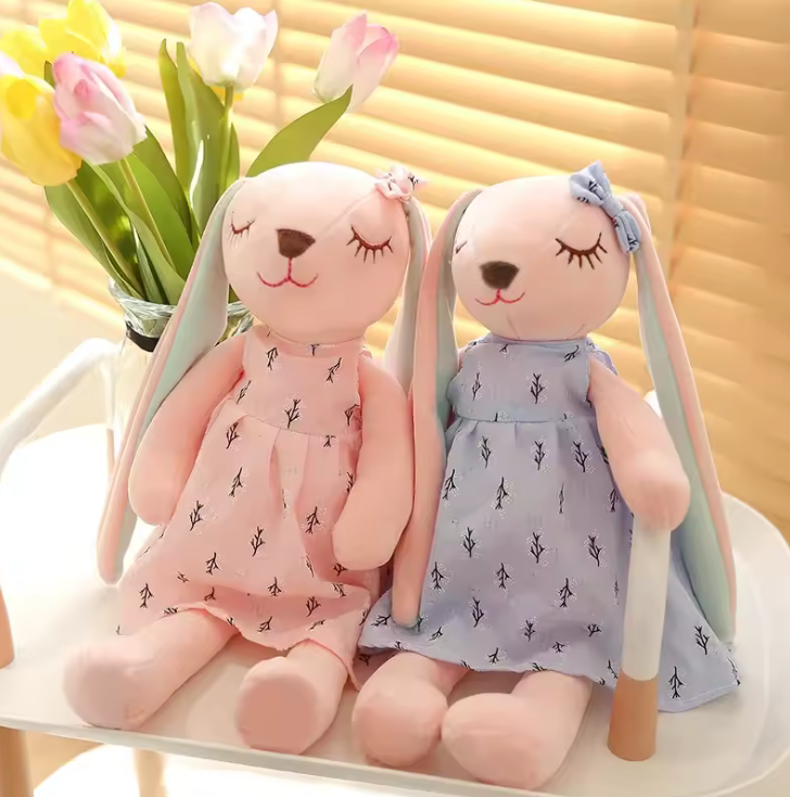 Peluche en forme de lapin rose portant une robe, avec de longues oreilles et un nœud sur la tête, assise sur fond blanc.